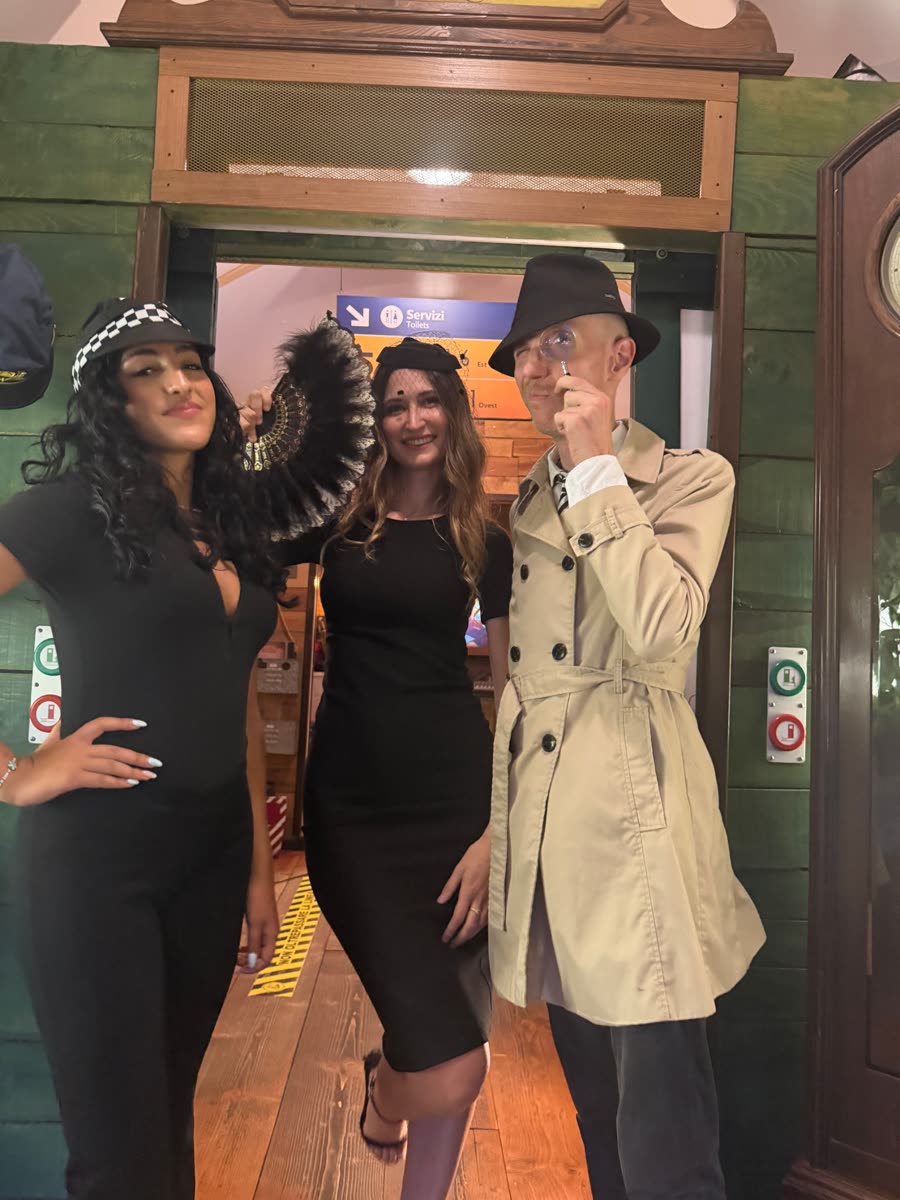 Tre persone in costume noir: berretto a scacchi, veletta e ventaglio, trench e lente d'ingrandimento